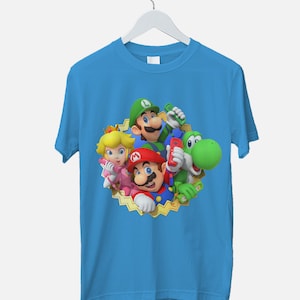 Peut inclure: Un t-shirt bleu clair avec un imprim&eacute; graphique de Mario, Luigi, Peach et Yoshi de la s&eacute;rie de jeux vid&eacute;o Super Mario. Les personnages sont repr&eacute;sent&eacute;s dans un cadre circulaire avec un motif d'&eacute;toile dor&eacute;e.