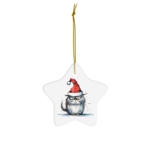 May include: A white star-shaped Christmas ornament featuring a grumpy cartoon cat wearing a red Santa hat. The cat has gray fur and an angry expression. A gold string is attached for hanging.