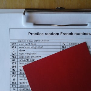 Könnte beinhalten: Ein weißer Clipboard mit einem roten Ordner darauf. Der Clipboard enthält ein Blatt Papier mit einer Liste französischer Zahlen und ihren schriftlichen Entsprechungen. Der Text auf dem Papier lautet "Practice random French numbers" und "Copyright © 2023 Sophie Chiasson".