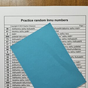 Könnte beinhalten: Ein weißes Blatt Papier, das teilweise von einem blauen Blatt Papier bedeckt ist. Das weiße Blatt enthält eine Liste von Zahlen und Innu-Wörtern. Der Text auf dem weißen Blatt lautet "Practice random Innu numbers" und "Copyright © 2023 Sophie Chiasson".