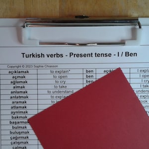 May include: A white clipboard with a red folder on top. The clipboard has a list of Turkish verbs in the present tense, with English translations. The verbs are written in black ink, and the translations are written in blue ink. The clipboard is on a wooden surface.