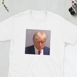 May include: White t-shirt featuring a color image of a man in a suit and red tie. The image appears to be a mugshot. The t-shirt is on a white surface with a camera, wallet, phone, and glasses.