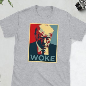 May include: A light gray t-shirt featuring a graphic of a man's portrait with the word "WOKE" in a rectangular box. The portrait has red, blue, and yellow tones. The t-shirt is displayed on a white surface.
