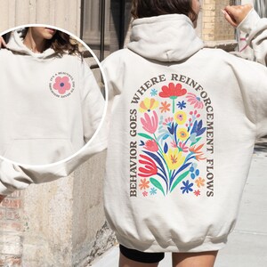 May include: Sand-colored hoodie with a floral design on the back and a smaller floral design on the front. The back design includes the text "WHERE REINFORCEMENT FLOWS" and "BEHAVIOR GOES". The front design includes the text "IT'S A BEAUTIFUL DAY TO SHAPE BEHAVIOR".