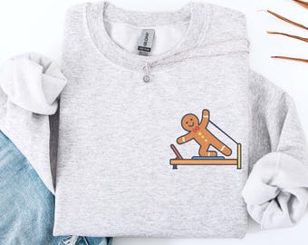May include: Light gray sweatshirt with a gingerbread man graphic doing Pilates. The gingerbread man is brown with white accents and is on a Pilates reformer. The word "ASH" is printed on the bottom left of the image.