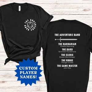 May include: Black t-shirt with white text and graphics. The front features a geometric design with the words "THE ADVENTURE BAND". The back lists character classes and names. A blue starburst graphic says "CUSTOM PLAYER NAMES!"
