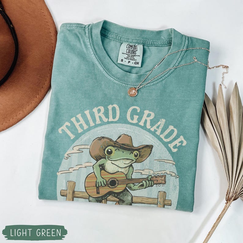 May include: Light green t-shirt with a graphic of a frog wearing a cowboy hat and playing a guitar. The text "THIRD GRADE" is printed above the frog. A silver necklace with a pendant is on the shirt.