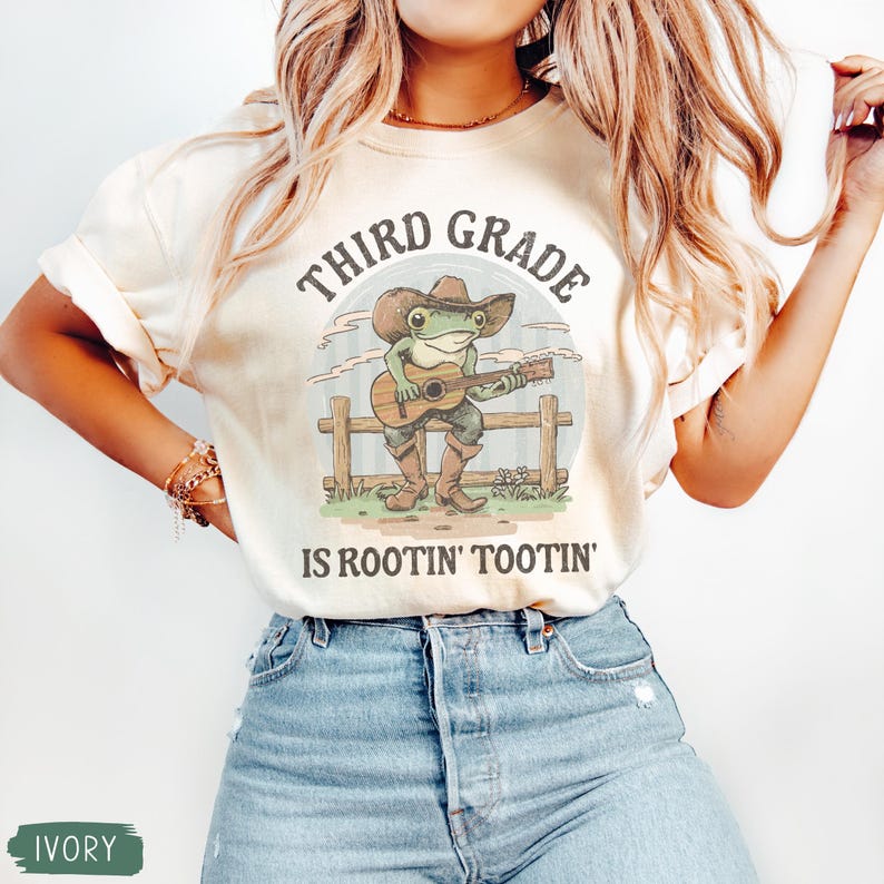 May include: Ivory-colored t-shirt featuring a cartoon frog playing a guitar, wearing a cowboy hat and boots. The text "THIRD GRADE" arches above the frog, with "IS ROOTIN' TOOTIN'" below. The design includes a fence and grassy area.
