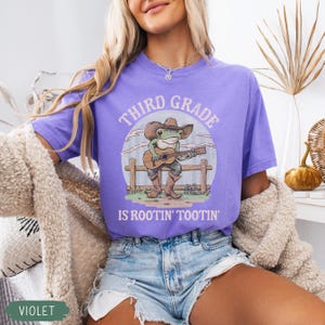 May include: Violet t-shirt with the text "Third Grade is Rootin' Tootin'". The shirt features a cartoon frog wearing a cowboy hat and boots, playing a guitar. The frog is set against a backdrop of a fence and a blue sky.