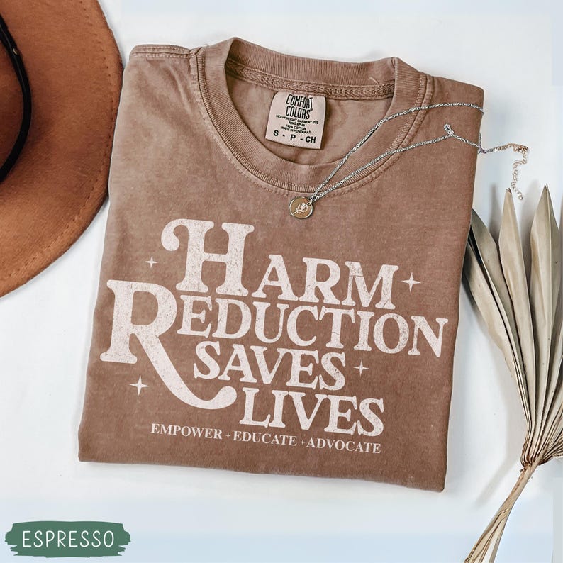 May include: Espresso-colored t-shirt with the text "Harm Reduction Saves Lives" in a white, stylized font. The shirt includes the phrases "Empower, Educate, Advocate". A silver necklace with a small pendant rests on the shirt.