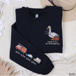 May include: Black long-sleeve shirt with a cartoon duck wearing a stethoscope and carrying a medical bag. The text "honk if it's an emergency" is below the duck. The left sleeve has illustrations of a fire truck, police car, and ambulance with the text "wee woo wee woo".