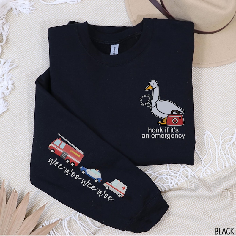 May include: Black long-sleeve shirt with a cartoon duck wearing a stethoscope and carrying a medical bag. The text "honk if it's an emergency" is below the duck. The left sleeve has illustrations of a fire truck, police car, and ambulance with the text "wee woo wee woo".