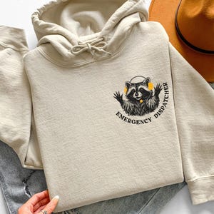 Peut inclure: Sweat à capuche beige avec cordon de serrage et un motif de raton laveur portant un casque et l'inscription "Emergency Dispatcher". Le sweat à capuche est plié sur un jean et un chapeau marron.