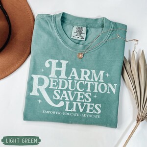May include: Light green t-shirt with the text "HARM REDUCTION SAVES LIVES" in white. The shirt also has the words "EMPOWER &bull; EDUCATE &bull; ADVOCATE" below the main text. A silver necklace with a pendant is on the shirt.