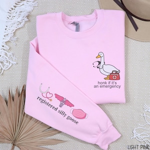 May include: Light pink sweatshirt with a cartoon goose wearing a stethoscope and carrying a medical bag. The text "honk if it's an emergency" is printed on the front. The left sleeve has the text "registered silly goose" with medical-themed graphics.