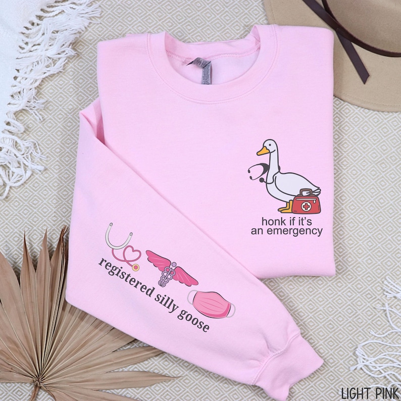 May include: Light pink sweatshirt with a cartoon goose wearing a stethoscope and carrying a medical bag. The text "honk if it's an emergency" is printed on the front. The left sleeve has the text "registered silly goose" with medical-themed graphics.