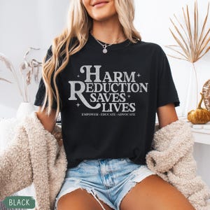 May include: Black t-shirt with the text "Harm Reduction Saves Lives" in a white, stylized font. The text is accompanied by the words "Empower, Educate, Advocate" below. The shirt is a casual style.