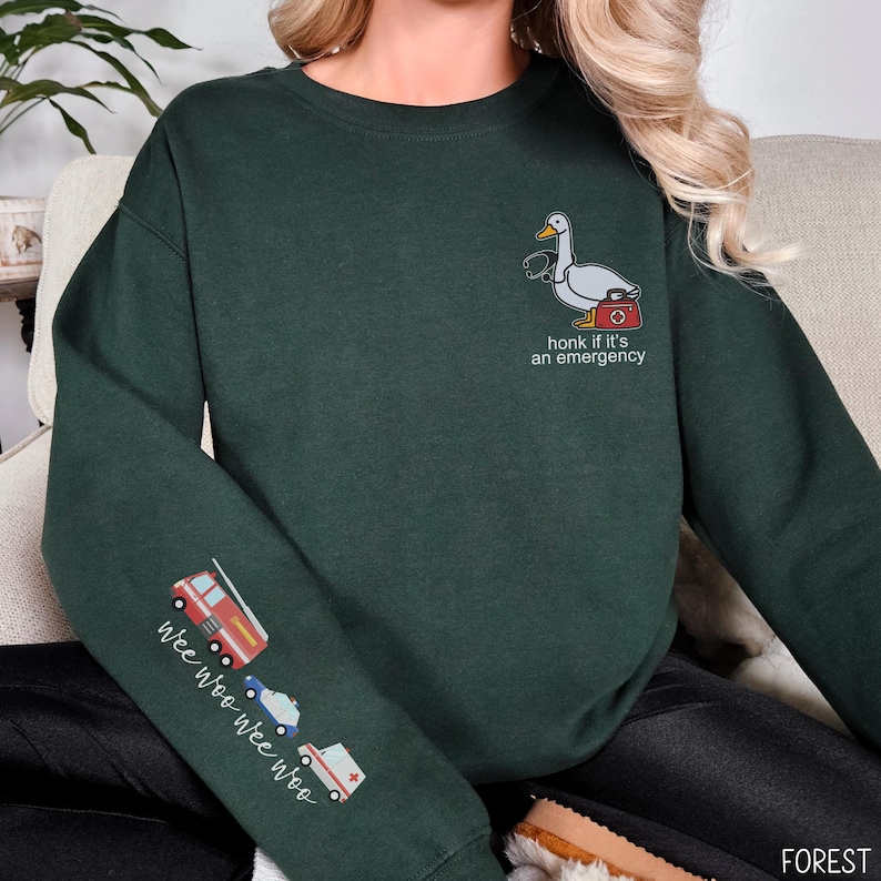 May include: Forest green sweatshirt with a cartoon goose wearing a stethoscope and carrying a medical bag. The text "honk if it's an emergency" is below the goose. The left sleeve has illustrations of emergency vehicles and the text "wee woo wee woo".
