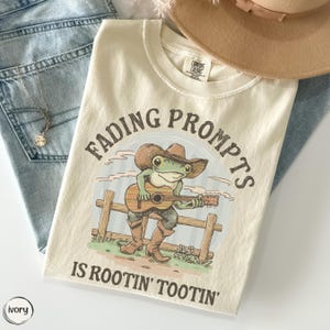 May include: An ivory t-shirt featuring a graphic of a green frog wearing a brown cowboy hat and boots, playing an acoustic guitar while sitting on a wooden fence. The text "FADING PROMPTS IS ROOTIN' TOOTIN'" is printed in dark brown lettering.