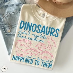 May include: An ivory t-shirt featuring blue text that reads "DINOSAURS didn't regulate their emotions and we know what HAPPENED TO THEM" alongside pink line-art illustrations of various dinosaurs including a T-Rex, triceratops, and pterodactyl.