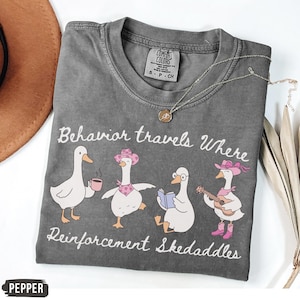 May include: A grey Comfort Colors t-shirt with the slogan "Behavior travels Where Reinforcement Skedaddles." The design shows four cartoon geese, each with a different accessory, including a coffee cup, cowboy hat, book, and guitar.