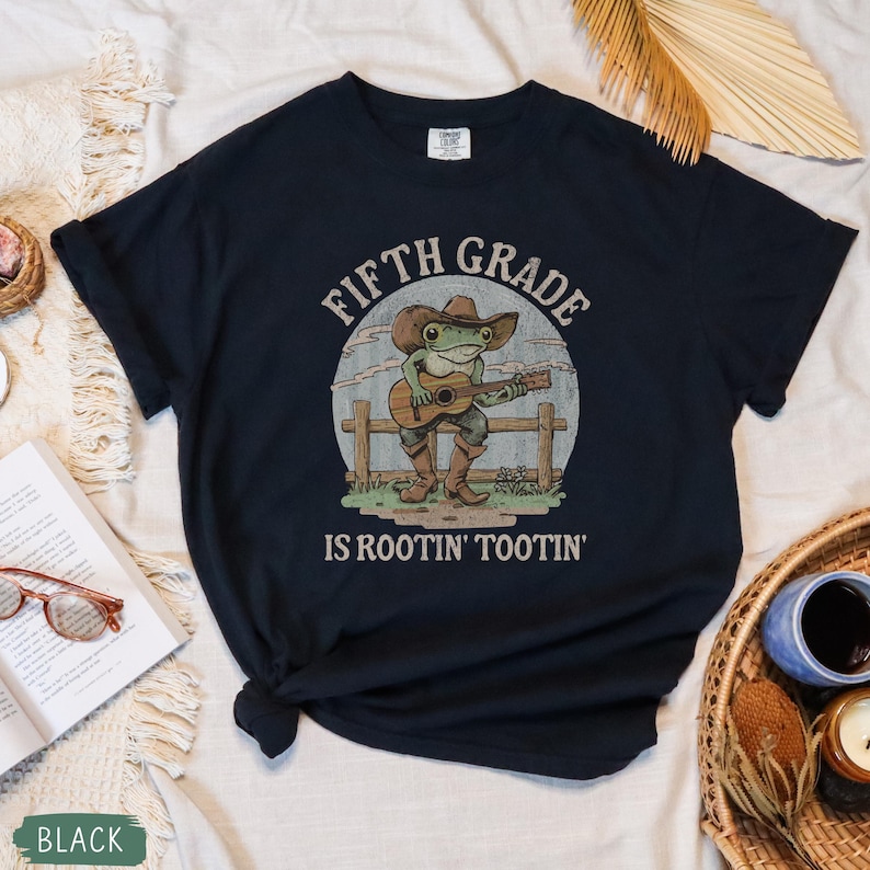 Puede incluir: Una camiseta negra de manga corta con el gr&aacute;fico de una rana verde con sombrero de vaquero y botas marrones tocando una guitarra ac&uacute;stica. El texto "FIFTH GRADE IS ROOTIN' TOOTIN'" aparece impreso en una fuente curva sobre y bajo la rana.