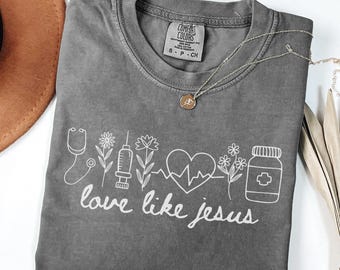 May include: A gray Comfort Colors t-shirt with a white graphic of medical symbols and flowers, along with the words "love like jesus." A silver necklace with a pendant is draped on the shirt.