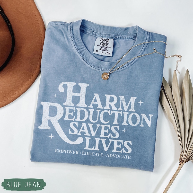 May include: A blue jean colored t-shirt with the text "HARM REDUCTION SAVES LIVES" in white. The shirt has a crew neck and short sleeves. A gold necklace with a pendant and a brown hat are also in the image.