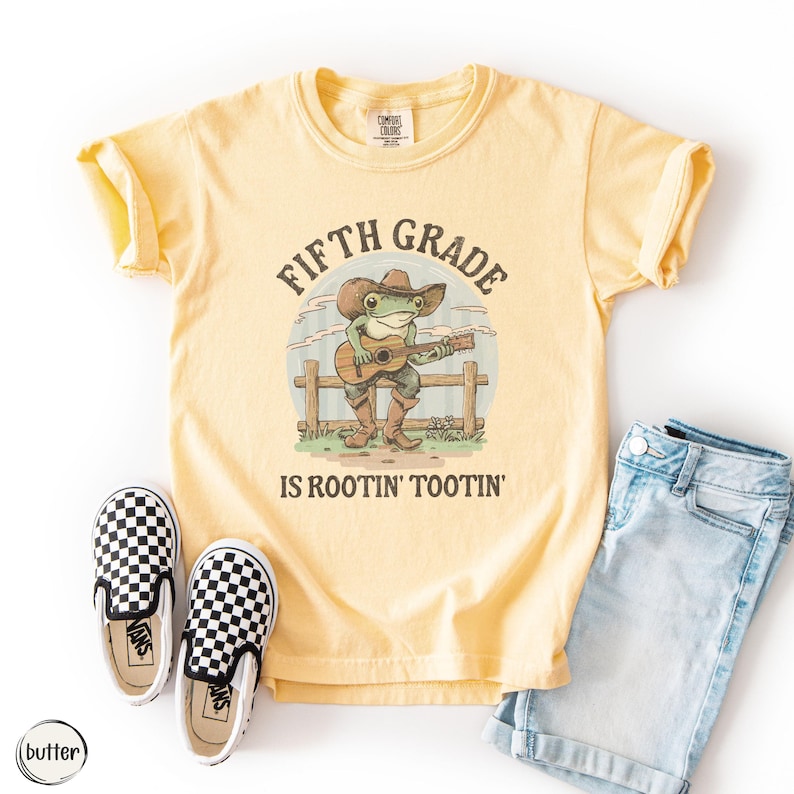 Puede incluir: Una camiseta de manga corta de color amarillo p&aacute;lido con el gr&aacute;fico de una rana con sombrero de vaquero y botas tocando una guitarra ac&uacute;stica sobre una valla de madera. El texto dice FIFTH GRADE IS ROOTIN' TOOTIN' en color marr&oacute;n.