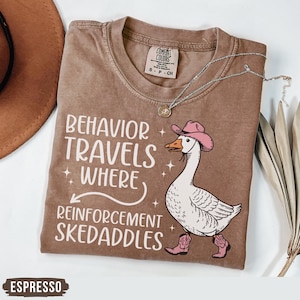 May include: Espresso-colored t-shirt with the phrase "Behavior Travels Where Reinforcement Skedaddles" and a cartoon goose wearing a pink cowboy hat and boots. A silver necklace is draped across the shirt.