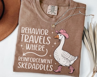 May include: Espresso-colored t-shirt with the phrase "Behavior Travels Where Reinforcement Skedaddles" and a cartoon goose wearing a pink cowboy hat and boots. A silver necklace is draped across the shirt.
