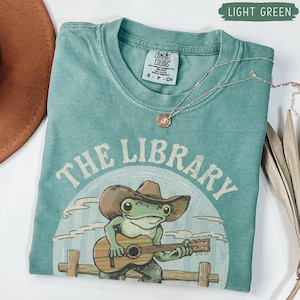 May include: Light green t-shirt featuring the text "THE LIBRARY" and a cartoon frog playing a guitar, wearing a cowboy hat. A silver necklace with a small pendant is draped on the shirt. The shirt is folded.