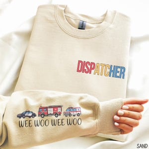 May include: A sand-coloured sweatshirt with the word "DISPATCHER" in colourful letters. The sleeve features illustrations of a police car, fire engine, and ambulance, with the text "WEE WOO WEE WOO" below. The sweatshirt is made of a soft material.