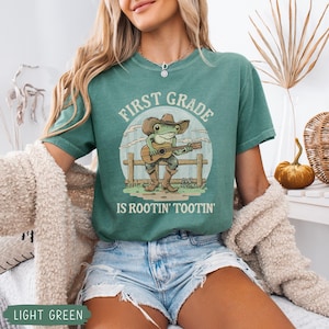 May include: Light green t-shirt with a graphic of a frog wearing a cowboy hat and boots, playing a guitar. The text on the shirt reads "FIRST GRADE IS ROOTIN' TOOTIN'".