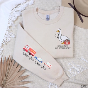 May include: Sand-colored sweatshirt with a cartoon duck wearing a stethoscope and carrying a medical bag. The text "honk if it's an emergency" is below the duck. The left sleeve has a fire truck, ambulance, and police car with the text "wee woo wee woo".