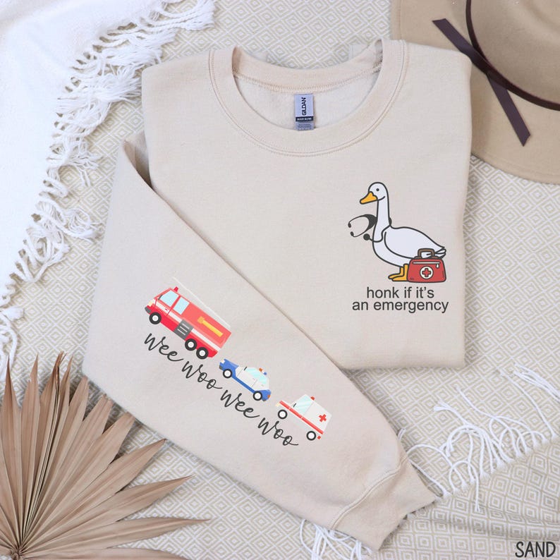 May include: Sand-colored sweatshirt with a cartoon duck wearing a stethoscope and carrying a medical bag. The text "honk if it's an emergency" is below the duck. The left sleeve has a fire truck, ambulance, and police car with the text "wee woo wee woo".