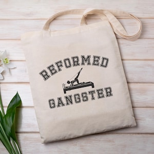 Pu&ograve; includere: Una borsa tote in tela bianca con testo nero che recita "REFORMED GANGSTER". Il testo &egrave; in un font universitario. Sopra il testo c'&egrave; una silhouette di una persona che fa un esercizio di pilates su un reformer.