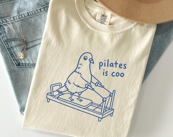 Può includere: Una t-shirt color avorio con un disegno a linee blu di un piccione che utilizza un macchinario per pilates. Il testo "pilates is coo" è stampato in blu accanto all'uccello. La t-shirt presenta un'etichetta del marchio Comfort Colors.
