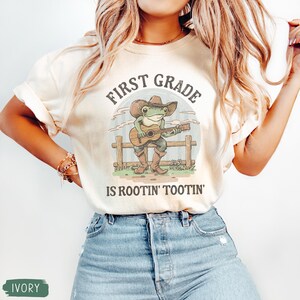 May include: Ivory-colored t-shirt with a graphic of a frog playing a guitar, wearing a cowboy hat and boots. The text reads "FIRST GRADE IS ROOTIN' TOOTIN'". The shirt is paired with blue jeans.