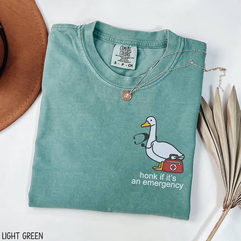 May include: Light green t-shirt with a cartoon goose wearing a stethoscope and carrying a first aid kit. The text "honk if it's an emergency" is printed below the goose.