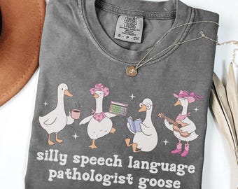May include: A grey Comfort Colors t-shirt with a playful design of four cartoon geese. Each goose is accessorised with items like a coffee cup, calculator, glasses, and a guitar. The text "silly speech language pathologist goose" is printed below the geese.