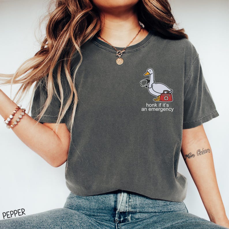 May include: A dark gray t-shirt with a white cartoon goose wearing a stethoscope and holding a first aid kit. The text "honk if it's an emergency" is printed below the goose.
