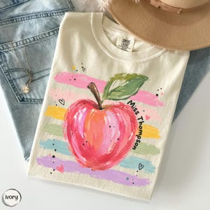 May include: An ivory t-shirt featuring a central graphic of a red apple with a green leaf, layered over horizontal pastel brushstrokes in pink, yellow, green, and purple. The text "Miss Thompson" is printed in black next to the apple.