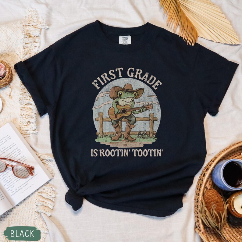 May include: Black t-shirt with a graphic of a frog wearing a cowboy hat and boots, playing a guitar. The text "FIRST GRADE" arches above the frog, and "IS ROOTIN' TOOTIN'" is below.
