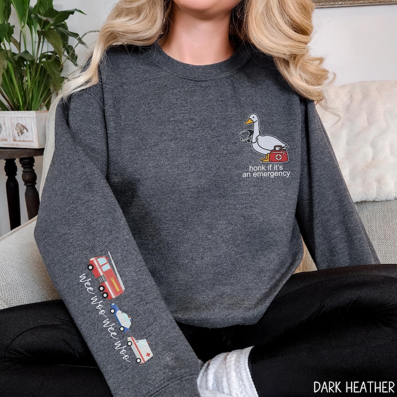 May include: Dark heather gray sweatshirt with a goose wearing a stethoscope and a medical bag, with the text "honk if it's an emergency." The left sleeve features a fire truck, police car, and ambulance with the text "wee woo wee woo."