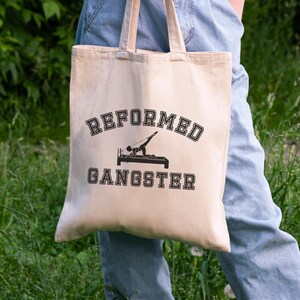 Pu&ograve; includere: Una borsa tote beige con testo nero che recita "REFORMED GANGSTER" e una silhouette di una persona che fa un esercizio di pilates.