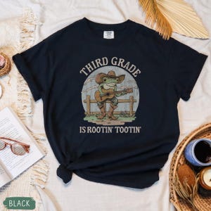 May include: Black t-shirt featuring a cartoon frog playing a guitar, wearing a cowboy hat and boots. The text on the shirt reads "THIRD GRADE IS ROOTIN' TOOTIN'". The design is set against a light blue and white background.