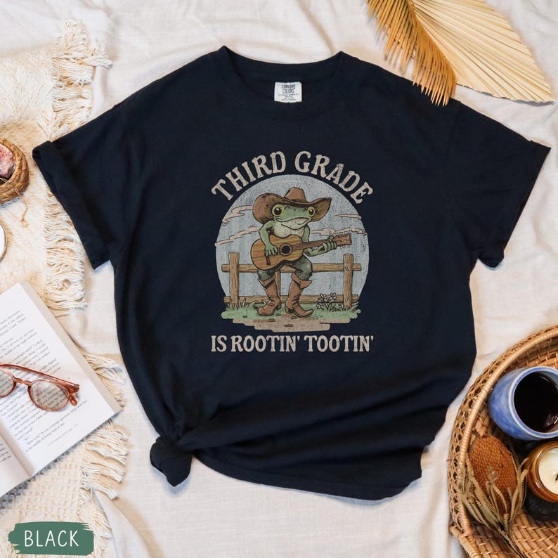 May include: Black t-shirt featuring a cartoon frog playing a guitar, wearing a cowboy hat and boots. The text on the shirt reads "THIRD GRADE IS ROOTIN' TOOTIN'". The design is set against a light blue and white background.