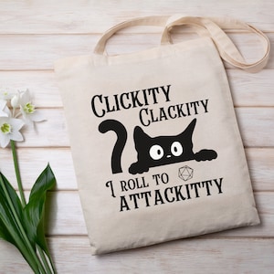 May include: Cream-coloured tote bag featuring a black cat illustration and text. The text says "Clickity Clackity, I Roll to Attackitty" with a D20 die. The bag has long straps and is made of canvas.