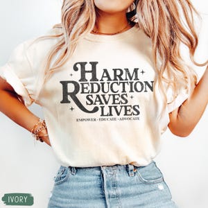 May include: Ivory-colored t-shirt with the text "Harm Reduction Saves Lives" in a bold, black font. The shirt also includes the words "Empower. Educate. Advocate." in a smaller font. The model is wearing blue jeans.